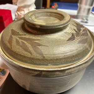 May include: A brown ceramic pot with a lid. The pot has a textured surface and a design of brown leaves on the side. The lid has a smooth surface.