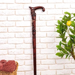 Puede incluir: Un bastón de madera de color marrón oscuro con un mango y un eje tallados de forma ornamentada con detalles florales. El bastón tiene un acabado liso y pulido y una punta de goma negra en la base, apoyado contra una pared de ladrillo blanco.