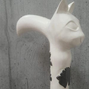 Pu&ograve; includere: Un bastone bianco a forma di gatto con un'impugnatura curva. La testa del gatto &egrave; in cima, con orecchie dettagliate e un'espressione serena. Il corpo del bastone &egrave; decorato con motivi a forma di foglia grigio scuro. Lo sfondo &egrave; un muro grigio.