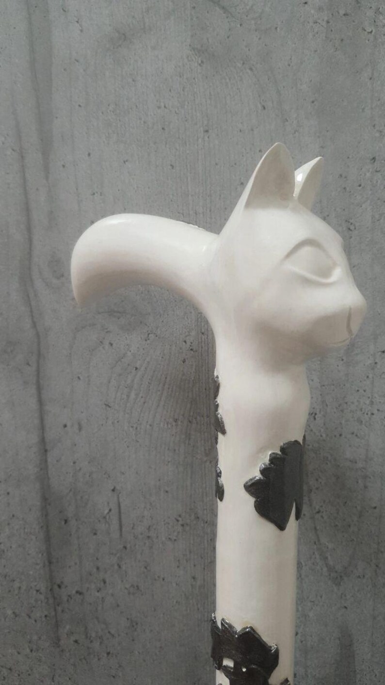 Pu&ograve; includere: Un bastone bianco a forma di gatto con un'impugnatura curva. La testa del gatto &egrave; in cima, con orecchie dettagliate e un'espressione serena. Il corpo del bastone &egrave; decorato con motivi a forma di foglia grigio scuro. Lo sfondo &egrave; un muro grigio.