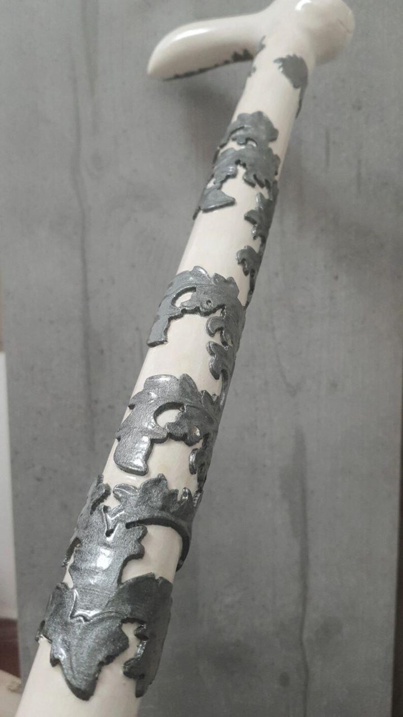 Pu&ograve; includere: Un bastone bianco con un'impugnatura curva e disegni argentati di rami d'albero che avvolgono l'asta. Il bastone &egrave; un oggetto decorativo, forse per la moda o il supporto.