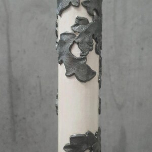 Pu&ograve; includere: Una candela color crema con decorazioni a foglie grigio scuro. Le foglie sono disposte in motivi verticali attorno alla forma cilindrica della candela. La candela &egrave; posta su uno sfondo grigio neutro.