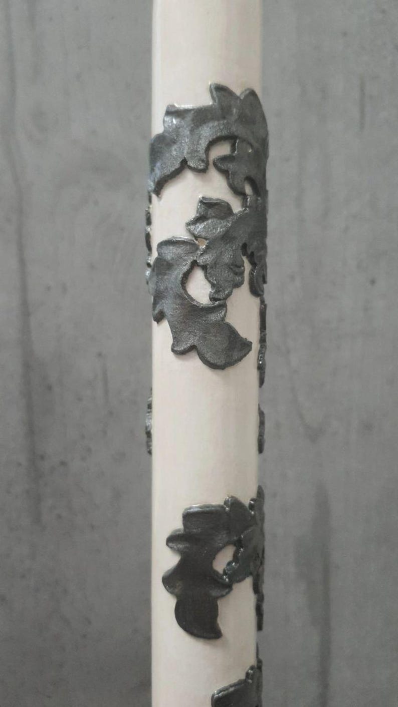 Pu&ograve; includere: Una candela color crema con decorazioni a foglie grigio scuro. Le foglie sono disposte in motivi verticali attorno alla forma cilindrica della candela. La candela &egrave; posta su uno sfondo grigio neutro.
