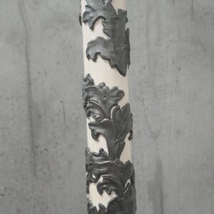 Pu&ograve; includere: Un oggetto decorativo cilindrico bianco con motivi a forma di foglia grigio scuro che avvolgono la sua superficie. Il design &egrave; in rilievo, creando un aspetto strutturato. L'oggetto &egrave; posto su uno sfondo grigio neutro.
