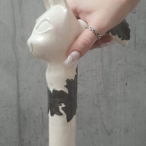 Pu&ograve; includere: Un bastone bianco a forma di gatto con un motivo decorativo a foglie grigio scuro. Il bastone &egrave; tenuto in mano con un anello d'argento. La testa del gatto &egrave; in cima al bastone.