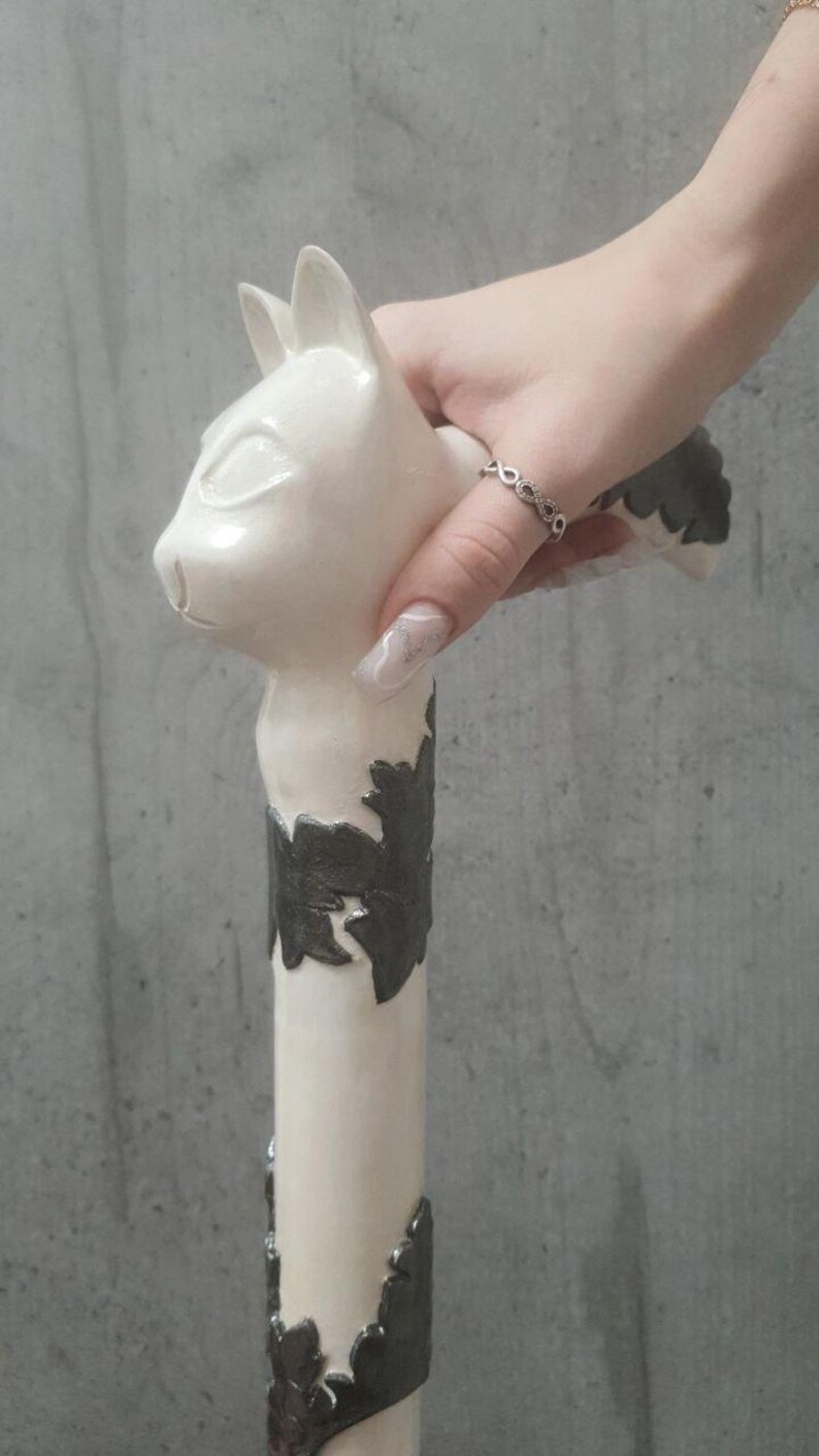 Pu&ograve; includere: Un bastone bianco a forma di gatto con un motivo decorativo a foglie grigio scuro. Il bastone &egrave; tenuto in mano con un anello d'argento. La testa del gatto &egrave; in cima al bastone.