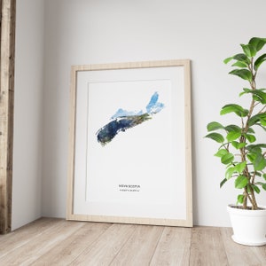 May include: A watercolor print of the outline of Nova Scotia, Canada. The print is framed in a light wood frame and is hanging on a white wall. The print is surrounded by a white mat.