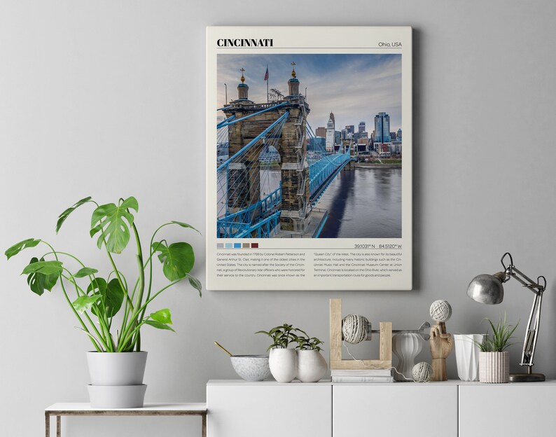 Może przedstawiać: Plakat w stylu vintage z fotografią mostu wiszącego Johna A. Roeblinga w Cincinnati, Ohio. Most jest stalową konstrukcją w kolorze niebieskim z brązową kamienną wieżą. Plakat zawiera nazwę miasta "Cincinnati" i stan "Ohio, USA" w minimalistycznym kroju pisma. Plakat zawiera r&oacute;wnież wsp&oacute;łrzędne "39.1031&deg;N, 84.5150&deg;W" oraz kr&oacute;tki opis miasta.