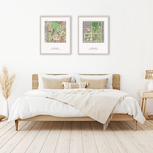 May include: Two framed maps of a university campus, hanging on a white wall above a bed with white bedding and a beige throw blanket. The bed has a wooden frame and two beige pillows. A vase with dried pampas grass is on the left side of the image, and a nightstand with a lamp is on the right side.