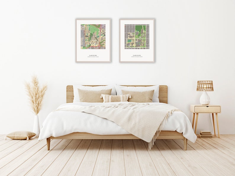May include: Two framed maps of a university campus, hanging on a white wall above a bed with white bedding and a beige throw blanket. The bed has a wooden frame and two beige pillows. A vase with dried pampas grass is on the left side of the image, and a nightstand with a lamp is on the right side.