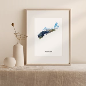 May include: A watercolor print of the outline of Nova Scotia, Canada, with a photo of the landscape overlaid on the map. The print is framed in a light wood frame and is hanging on a light beige wall.