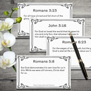 May include: Four white cards with black text and grey borders. The cards feature Bible verses from Romans and John. The text on the cards reads: "Romans 3:23 For all have sinned and fall short of the glory", "John 3:16 For God so loved the world that he gave his one and only Son, that whoever believes in him shall not", "Romans 6:23 For the wages of sin is death, but the gift of God is eternal life in Christ Jesus our Lord.", and "Romans 5:8 But God demonstrates his own love for us in this: While we were still sinners, Christ died for us." The cards are on a wooden surface with a black pen.
