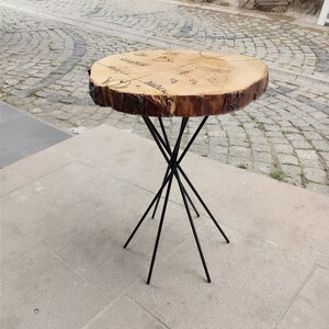 Puede incluir: Una mesa redonda de madera con una tapa de madera natural y patas de metal negro. La tapa de la mesa tiene un dise&ntilde;o &uacute;nico con un patr&oacute;n circular y las palabras "Naturaleza" y "Madera" escritas en ella.