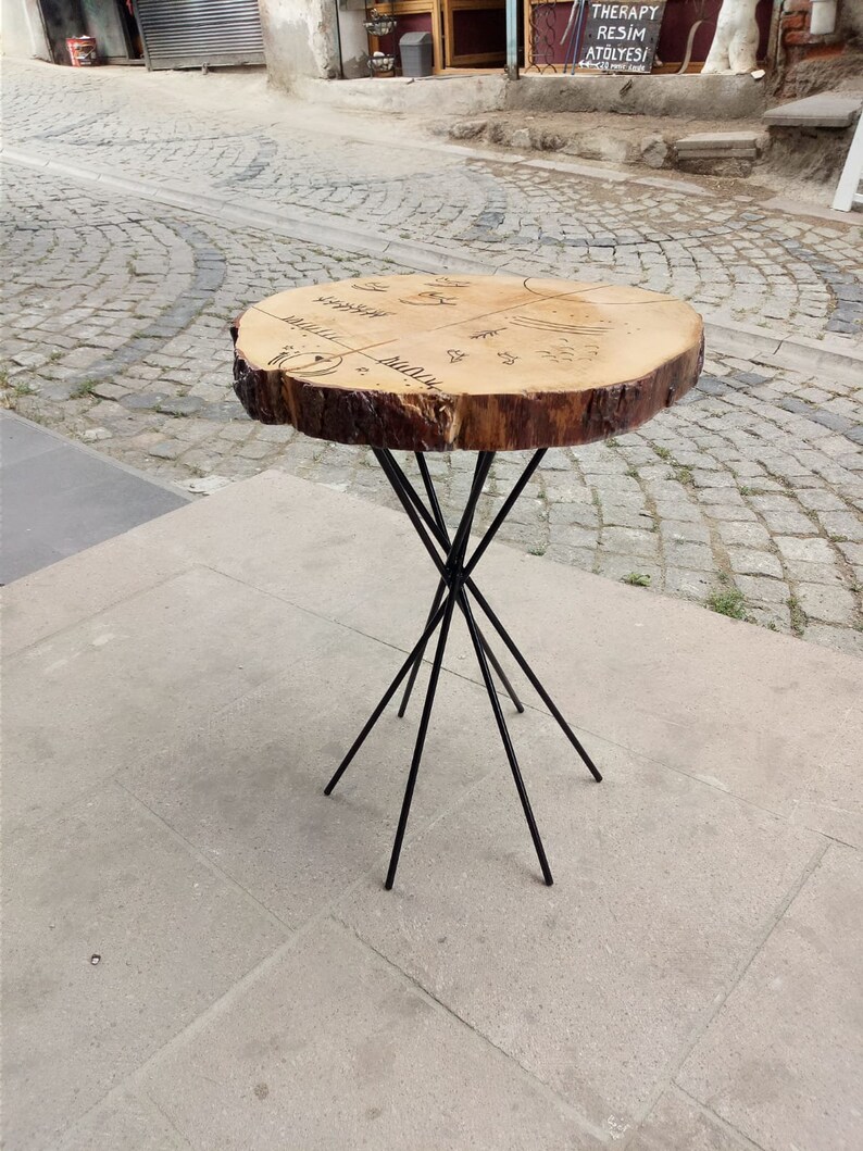 Puede incluir: Una mesa redonda de madera con una tapa de madera natural y patas de metal negro. La tapa de la mesa tiene un dise&ntilde;o &uacute;nico con un patr&oacute;n circular y las palabras "Naturaleza" y "Madera" escritas en ella.