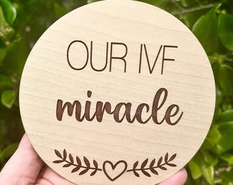 May include: A round wooden sign with the text "OUR IVF miracle" in brown lettering. The sign is decorated with a small heart and leafy branches.