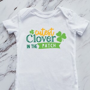 May include: White baby onesie with the text "cutest Clover in the Patch" in green and yellow. The design includes a shamrock. The onesie is a short-sleeved garment with snap closures.