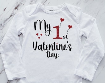May include: White long-sleeved baby onesie with the text "My 1st Valentine's Day" in black with a red glitter 1 and red heart accents.