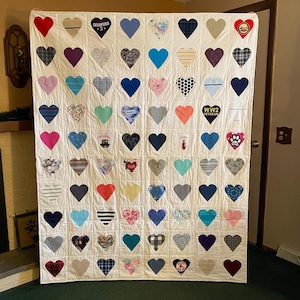 May include: A handmade quilt featuring a grid of heart-shaped patches in various colors and patterns, including solid colors, stripes, and floral prints. The quilt is displayed against a neutral background, showcasing the vibrant heart designs.