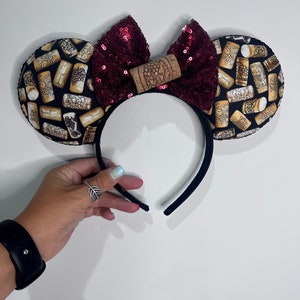 May include: A black headband with two round ears. The ears are patterned with wine corks. A sequined burgundy bow is centered on the headband, with a wine cork in the middle. The headband is designed to look like mouse ears.