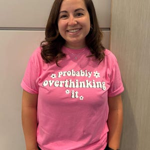 May include: A pink t-shirt with the phrase "probably overthinking it" in white, stylized text with daisy accents. The shirt is short-sleeved and worn by a person.