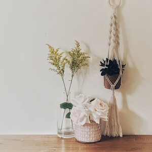 May include: A white macrame plant hanger with a small potted plant hanging from it. The hanger is hanging on a white wall. There are two vases with yellow flowers and white flowers in front of the hanger.