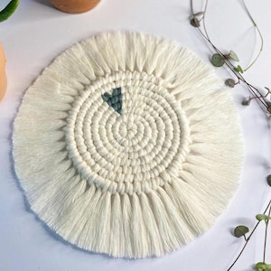 May include: A circular, cream-coloured macrame coaster with a fringed border. The centre has a spiral pattern and a teal heart detail. This handmade coaster is suitable for home use.