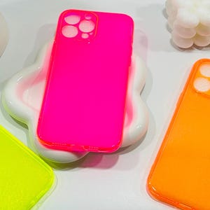 Puede incluir: Tres fundas de teléfono de neón en rosa, amarillo y naranja. Las fundas son de plástico transparente y tienen un acabado brillante.
