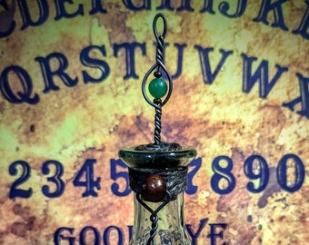 May include: A green glass bottle with a textured surface and a metal top with a green gemstone. The bottle is sitting on a brown surface in front of a vintage Ouija board.