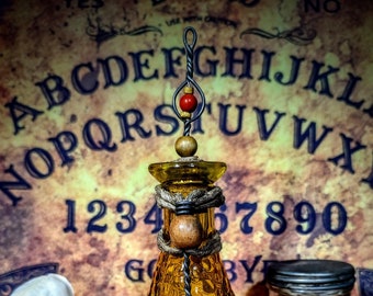 May include: A yellow glass bottle with a textured surface and a metal lid. The bottle is decorated with a wooden bead and a red bead. The bottle is sitting on a brown surface in front of a vintage Ouija board.