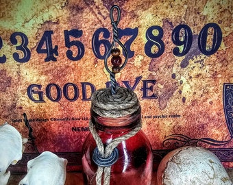 May include: A red glass bottle with a cork stopper and a rope tied around the neck. The bottle is sitting on a wooden surface with two animal skulls and a Ouija board in the background. The Ouija board has the numbers 1 through 9 and the letters GOODBYE.