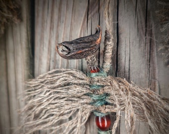May include: A hanging ornament crafted from natural fibres, featuring a dark, curved seed pod at the top resembling a creature's head. The body is wrapped in tan and teal twine with frayed edges, accented by two smooth, polished dark red beads.
