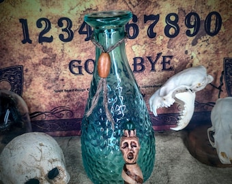 May include: A teal blue glass bottle with a cork stopper and a wooden figure with a face carved into it. The bottle is decorated with a brown rope and a wooden bead. The bottle is sitting on a brown surface in front of a vintage Ouija board.