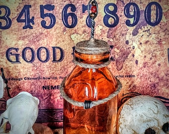 May include: A brown glass bottle with a cork stopper and a metal wire with red beads hanging from it. The bottle is tied with a brown rope and is sitting on a brown surface. The bottle is in front of a vintage Ouija board with the numbers 3, 4, 5, 6, 8, 9, 0 and the word "GOOD" visible.