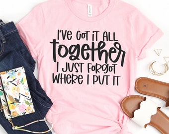 May include: A light pink t-shirt with the text "I've got it all together, I just forgot where I put it" in black. The shirt is styled with blue jeans, a floral wallet, and sandals.