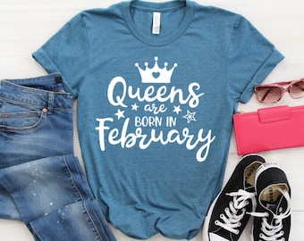 Puede incluir: Una camiseta azul turquesa con un diseño gráfico blanco que dice "Queens are born in February" con una corona y estrellas.