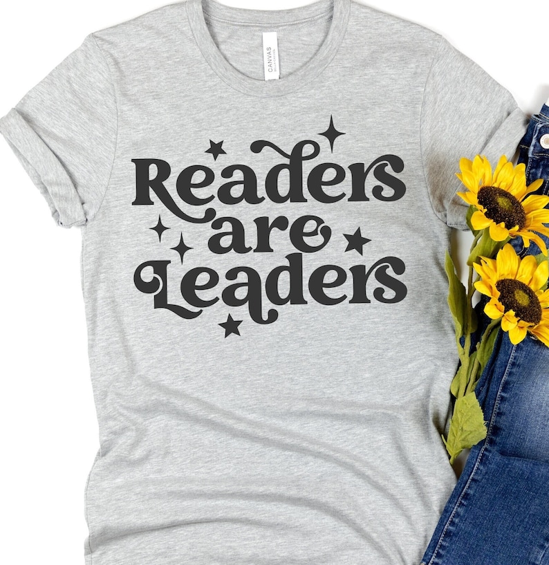 Pode incluir: Camiseta cinza com o texto "Readers are Leaders" em preto com estrelas ao redor do texto.