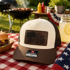 May include: A trucker hat with a cream-colored front panel, a brown brim, and a brown mesh back. A rectangular brown patch on the front features a graphic of an American flag and the number 250. A black sticker on the brim reads Richardson 112.