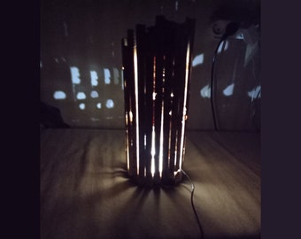 May include: A wooden lamp with a cylindrical shape, made of thin strips of wood, lit from within, casting a warm glow on the surface below.