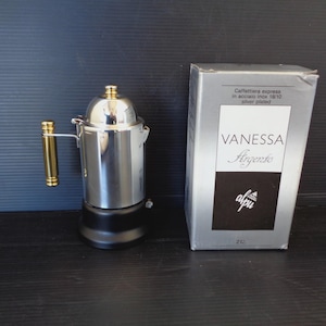 May include: A silver-toned stainless steel stovetop espresso maker with a black base and a gold-colored handle. Next to it is a silver box labeled Vanessa Argento, Caffettiera express in acciaio inox 18/10 silver plated, and the brand name alpu.