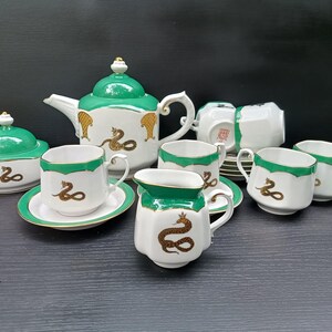 Może przedstawiać: Biały zestaw do herbaty z porcelany z zielonym obrzeżem i złotymi motywami węża. Zestaw zawiera dzbanek do herbaty z pokrywką, cukiernicę z pokrywką, dzbanek na mleko oraz kilka filiżanek ze spodkami, ozdobione wzorem złotego węża i korony.