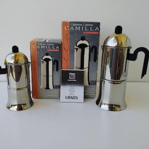 Könnte beinhalten: Zwei Espressokocher aus poliertem Edelstahl mit schwarzen Griffen und Knöpfen, begleitet von ihren originalen Pappverpackungen mit der Aufschrift Camilla Lavazza und einem kleinen Informationsheft mit dem aufgedruckten Lavazza-Logo.