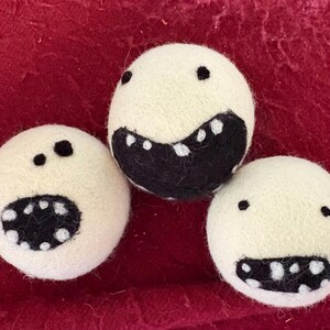 May include: Three off-white felt balls with black and white facial features. Each ball has a different expression, with black dots for eyes and black and white mouths. The balls are arranged on a red surface.