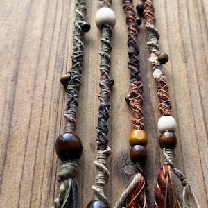 Op de afbeelding: Vier haarwraps met veelkleurige draad, garen en koord in aardse tinten bruin, groen en oranje. Elke wrap is versierd met diverse houten, metalen en stenen kralen van verschillende groottes, eindigend in losse, bungelende franjes aan de onderkant.