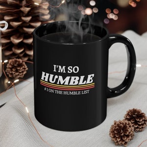 May include: Black coffee mug with white text that reads "I'm so humble #1 on the humble list" with a retro rainbow design.