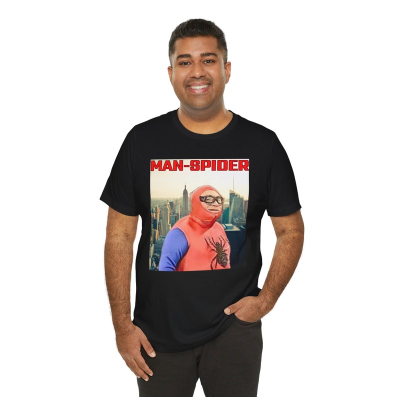 May include: Black t-shirt featuring a graphic of a person in a red and blue superhero costume with the text "MAN-SPIDER" in red letters. The superhero is wearing glasses and a hood, with a cityscape background.