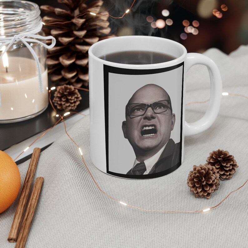 May include: White ceramic coffee mug featuring a black and white image of a man wearing glasses and a suit. He has a serious expression.