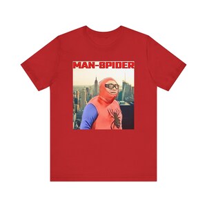 May include: Red t-shirt featuring a humorous image of a person in a homemade Spider-Man costume with the text "MAN-SPIDER" in a red box. The image shows a cityscape in the background.