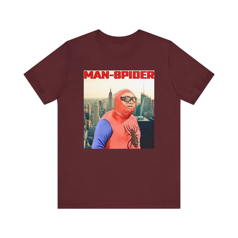 May include: A maroon t-shirt featuring a humorous image of a person dressed as a superhero, with the text "MAN-SPIDER" in a red box. The image shows a person in a red and blue costume with a spider graphic, set against a cityscape backdrop.