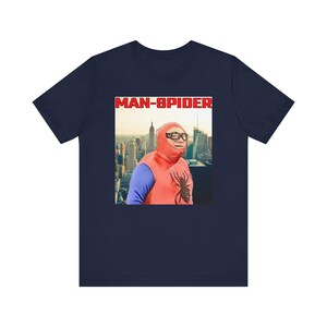 May include: Navy blue t-shirt featuring a humorous image of a person dressed as a superhero with the text "MAN-SPIDER" in red letters. The image shows a cityscape in the background. The superhero costume is red and blue.