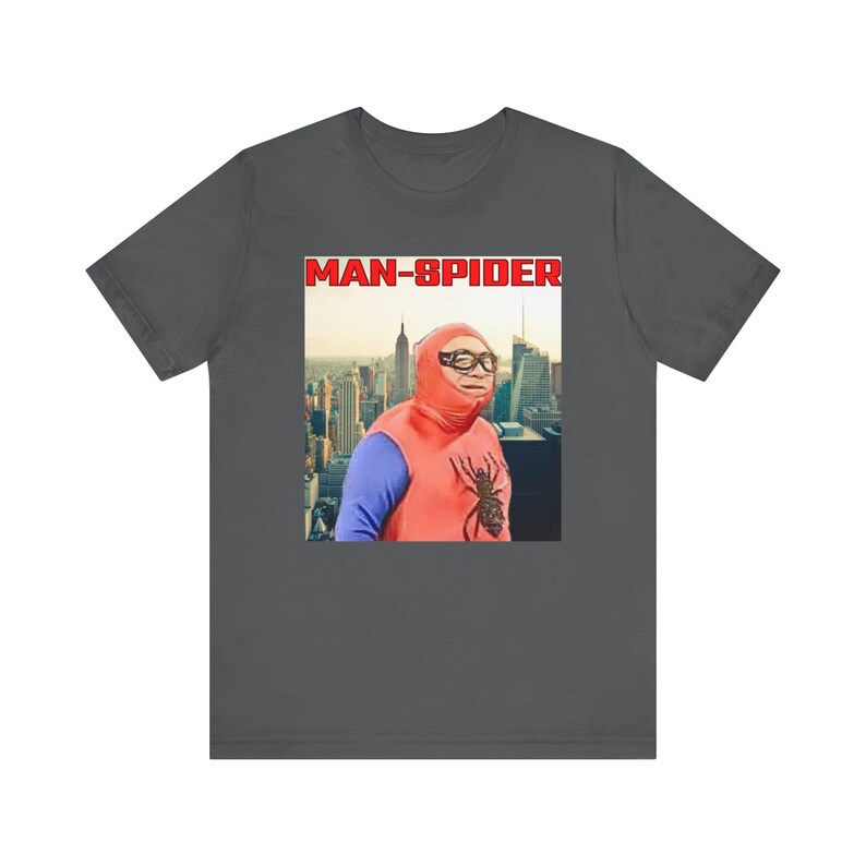 May include: Dark gray t-shirt featuring a graphic of a person in a red and blue costume with a spider emblem, and the text "MAN-SPIDER" in a red box. The background shows a cityscape.